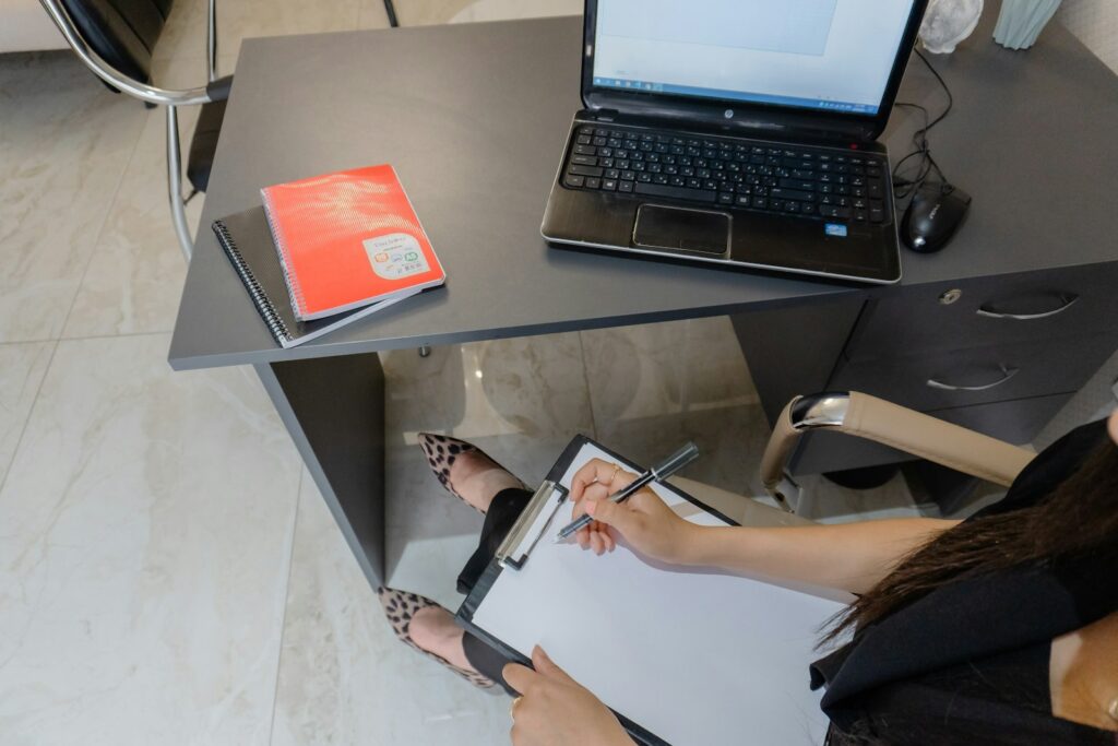 Pessoa preenchendo um formulário em prancheta ao lado de um notebook