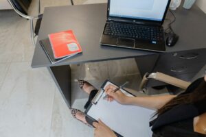 Pessoa preenchendo um formulário em prancheta ao lado de um notebook